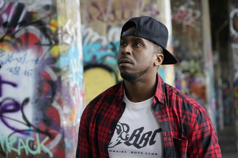 man with red shirt, black hat, and white tee - shirt standing next to a bunch of graffiti