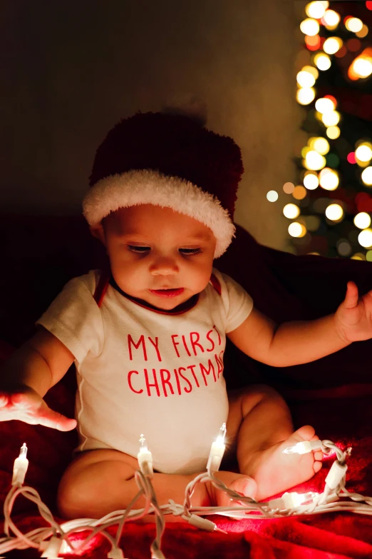 a little baby wearing a white shirt sitting next to a christmas tree