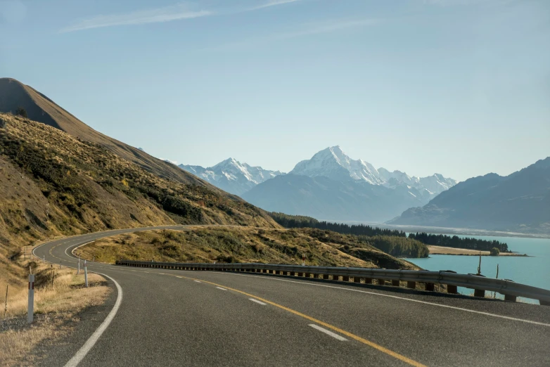 an empty highway leads into the mountains next to a lake