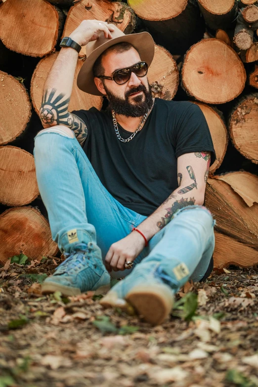 a man with tattoos sits on the ground near a pile of logs