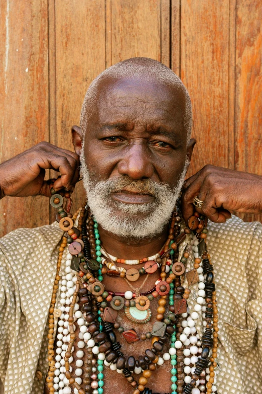 an older man in necklaces stands next to wooden wall