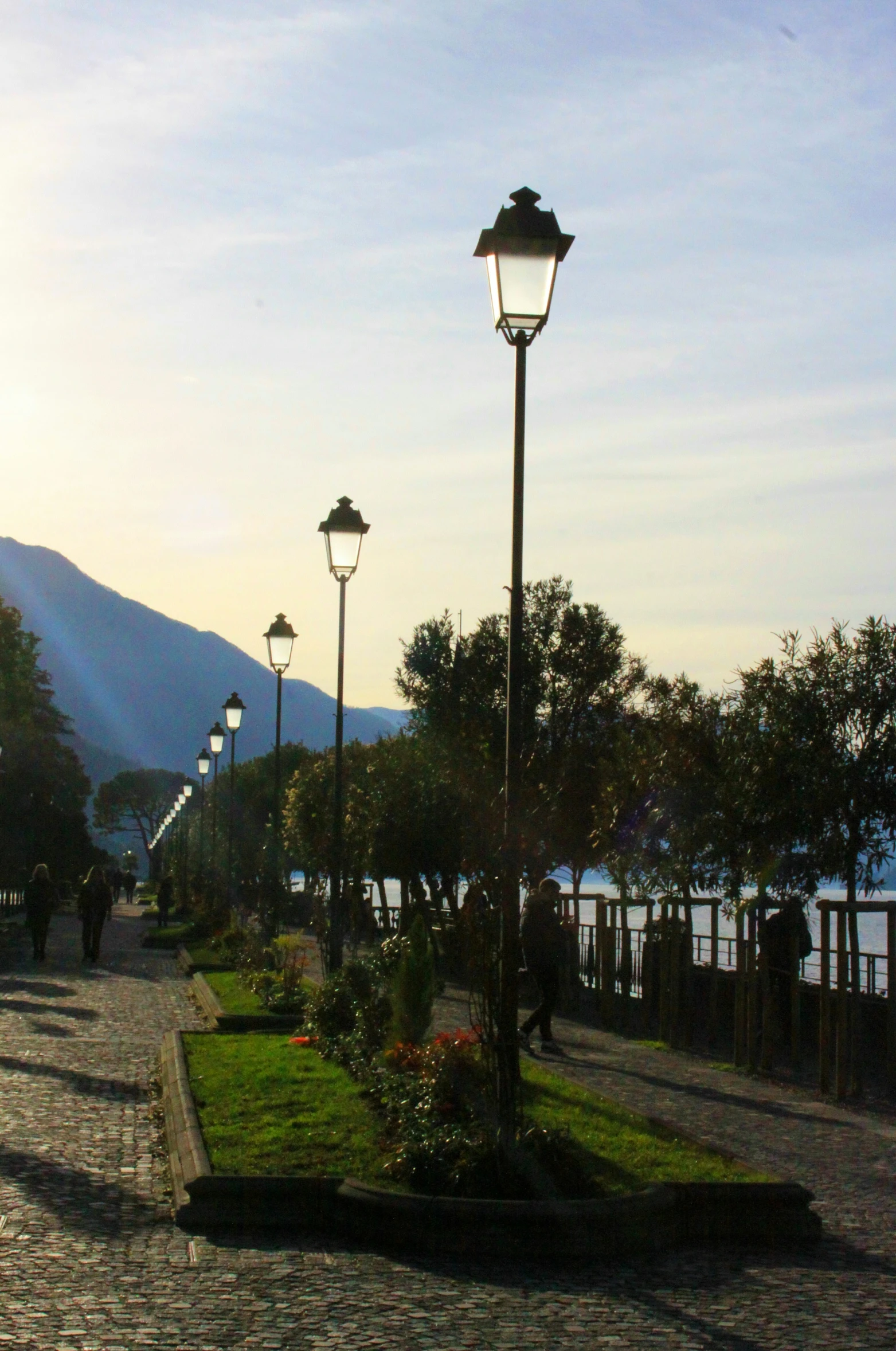 a brick path leading to street lights and flower beds in front of the water