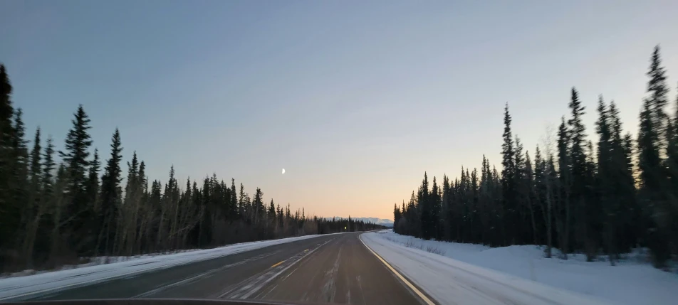 the sun is setting on a highway with snow and trees