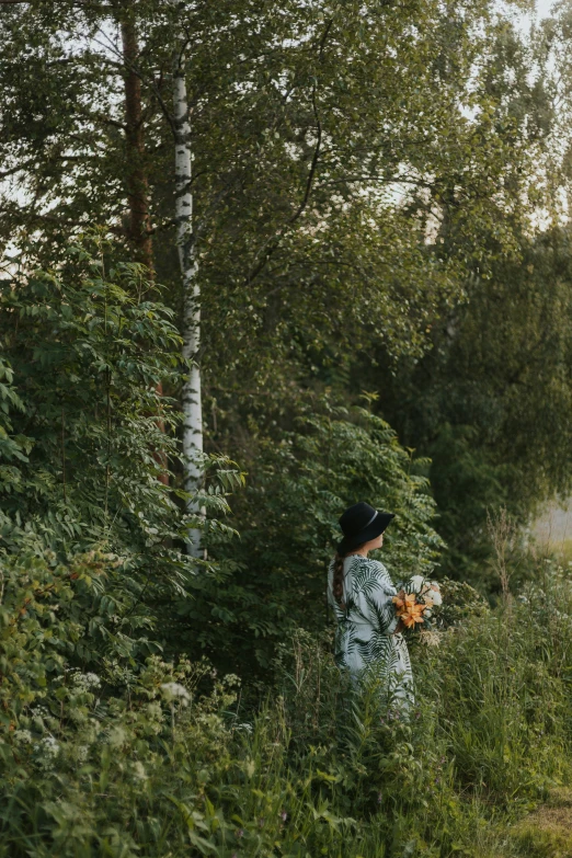 a woman with black hat walking through tall grass and bushes