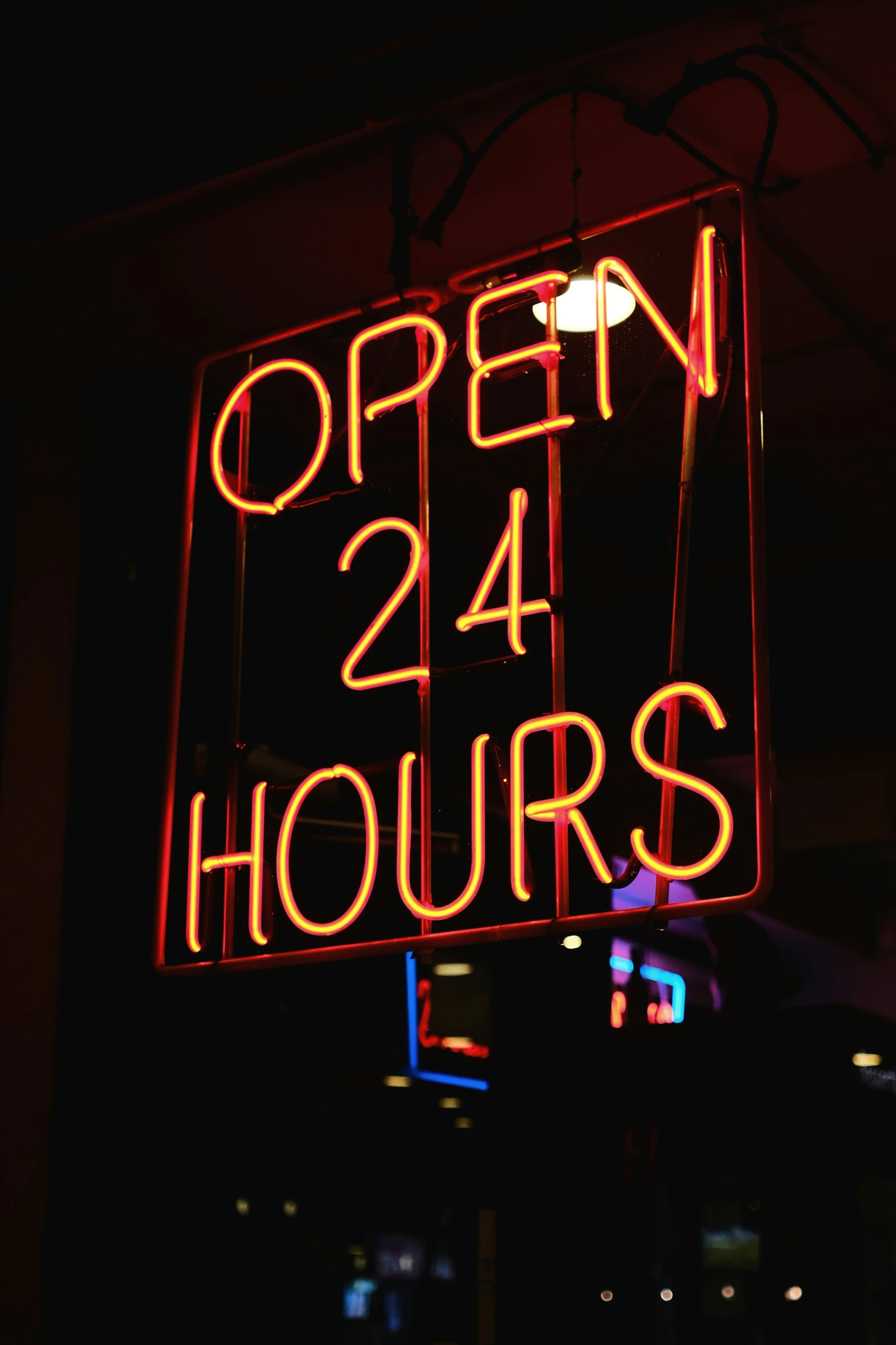 open 24 hours neon sign with city lights behind it