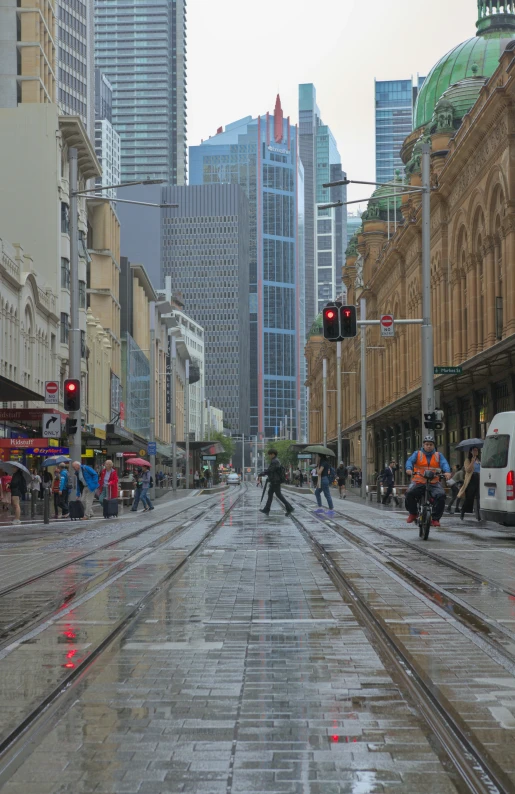 a wet city street with buildings and people sitting on a bench on the side of the track