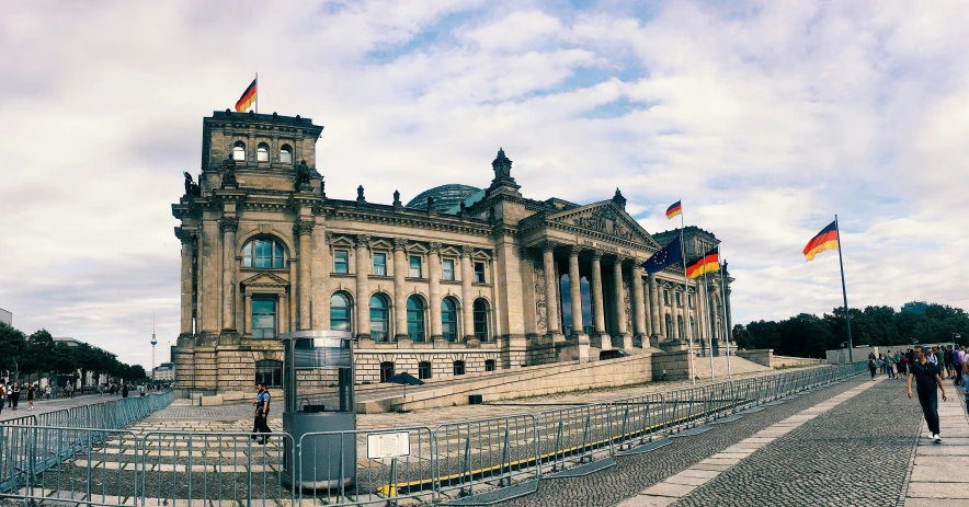 people walk on the cobblestone street near a large building with two flags flying