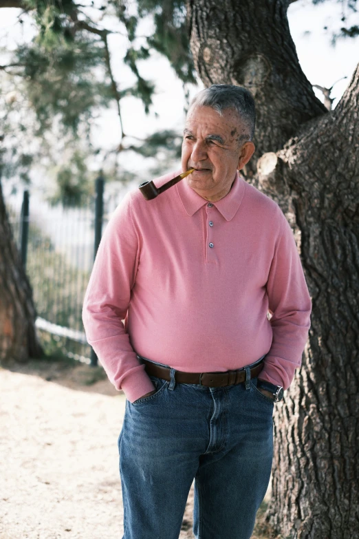 an old man in a pink shirt and blue jeans is leaning up against a tree