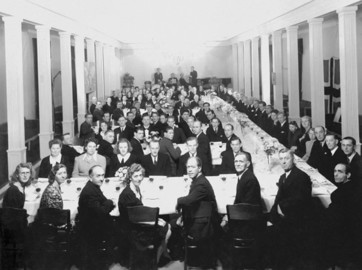 an old pograph of people sitting at tables with place settings in a hall