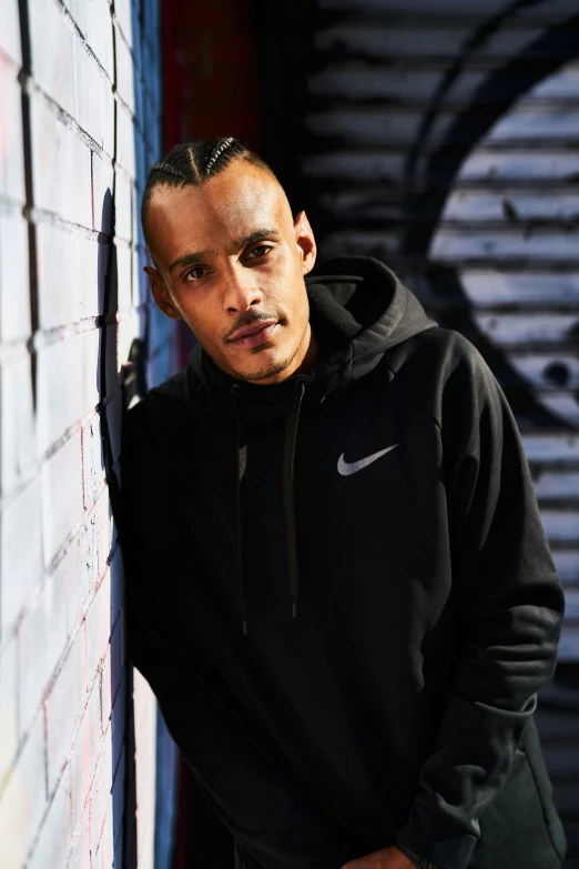 a young man in a black hoodie and sunglasses stands by a graffiti - covered wall