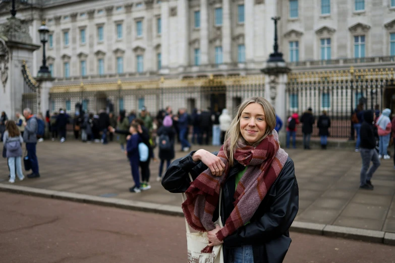 woman with brown hair in black jacket and red scarf in front of a building