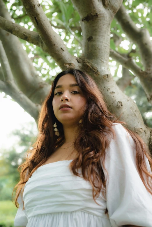 young woman wearing a white top poses by a tree