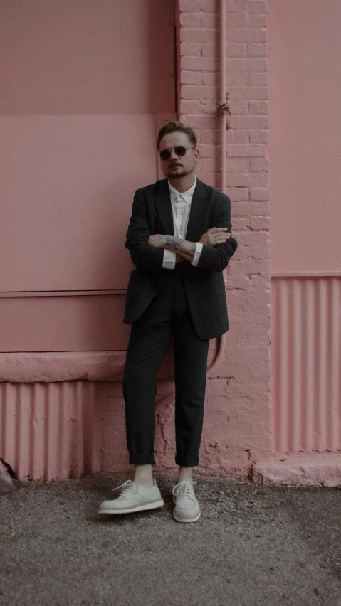 a man standing with his arms crossed leaning against a pink wall