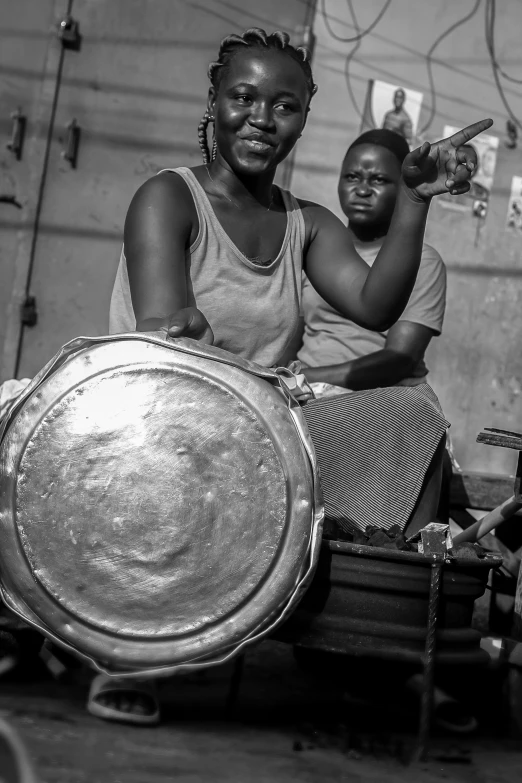 two woman sit with different sized drums while pointing