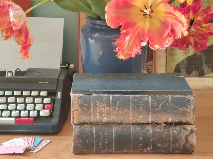 a pair of books, a typewriter, and flowers sit on a wooden table