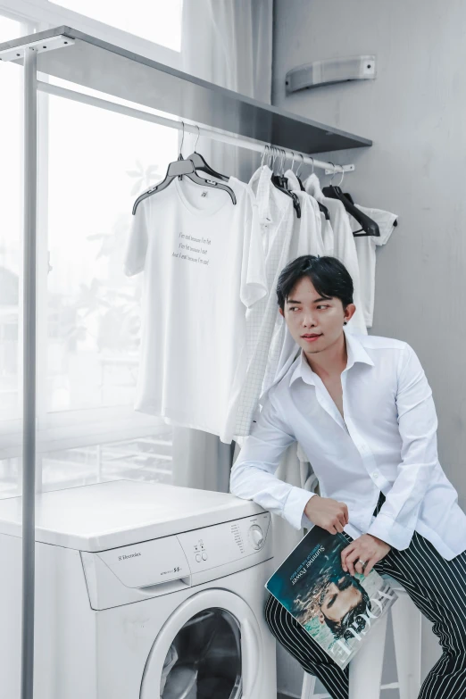 young man holding his book while sitting on the clothes rack beside a washer