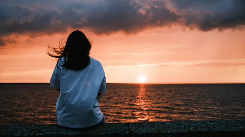 a woman sitting on the edge of the cliff watching the sun set