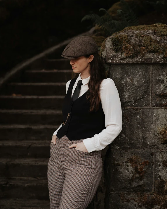 young woman leaning against a stone wall wearing suit and hat