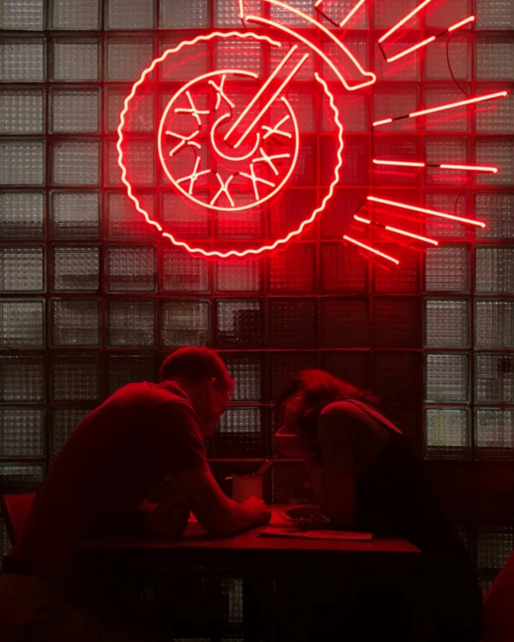 two people sitting down at a table talking in front of a wall with large red neon signs