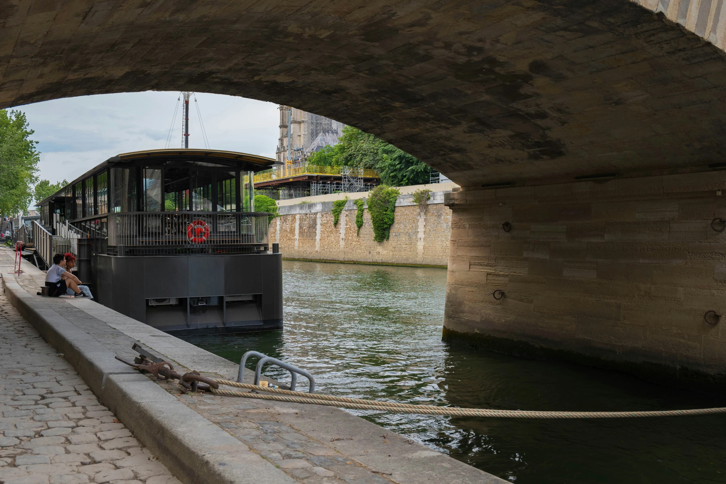 a large river boat traveling under a bridge next to a sidewalk