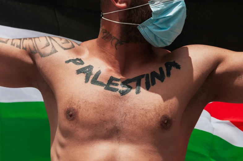 a man with a face mask wearing a shirt that says palestine on his chest and a green and white striped flag behind him