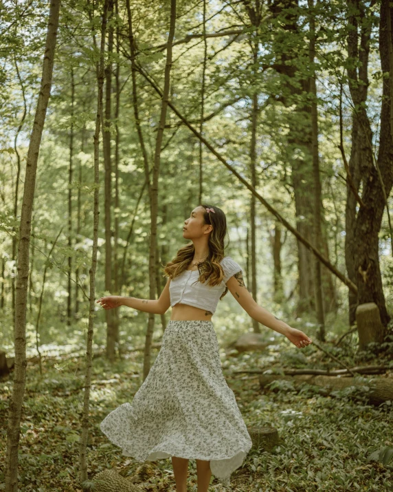 a woman in a white top standing in a wooded area with her hands extended