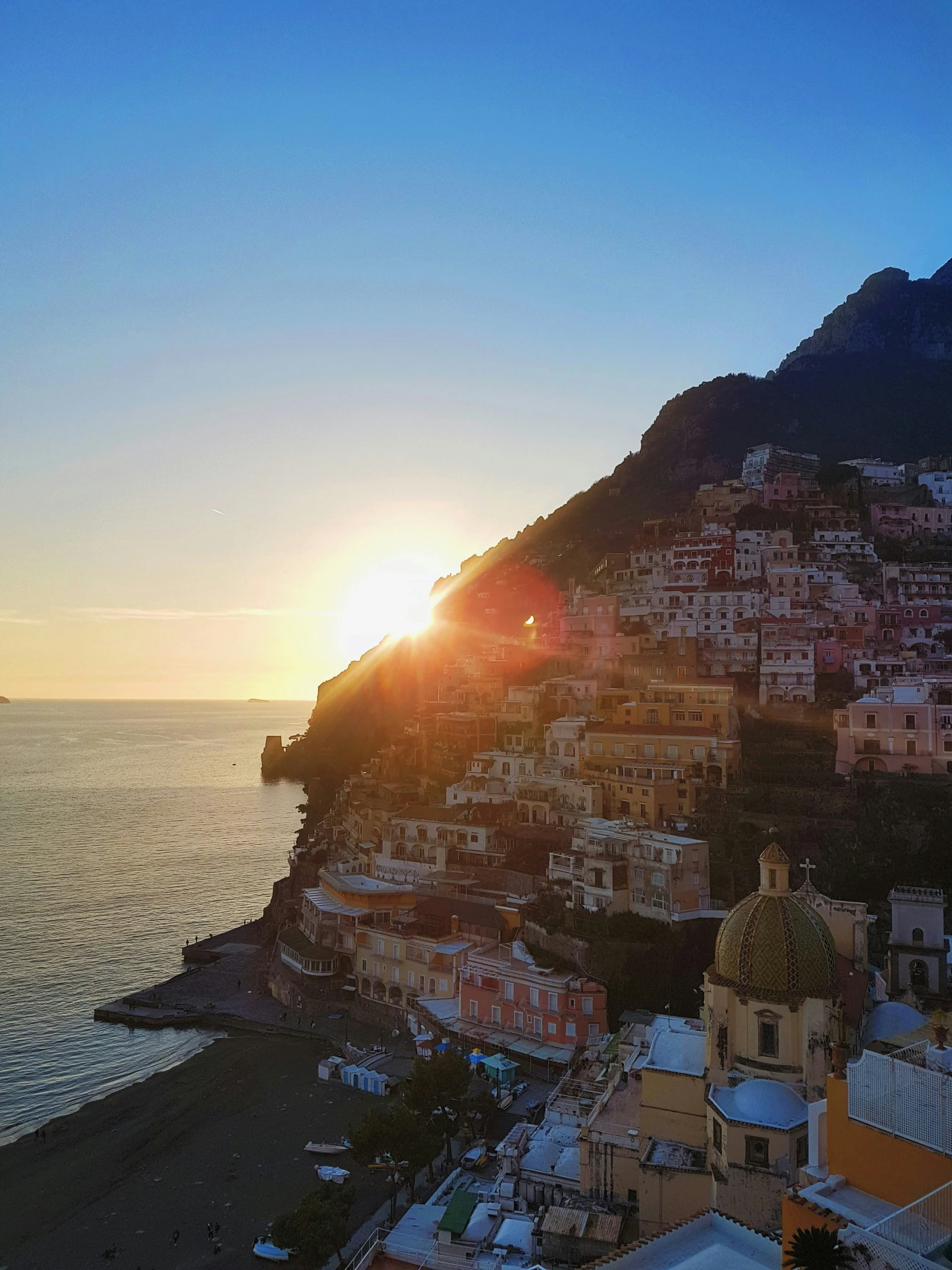 an aerial s shows the town of amalfata, in the distance, as the sun sets over the ocean