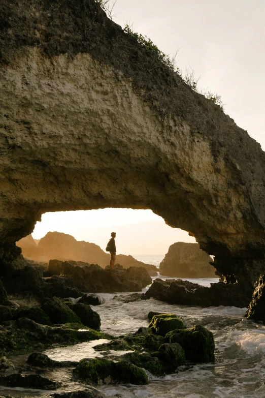 a rock formation sitting on the shoreline near a man standing in the ocean