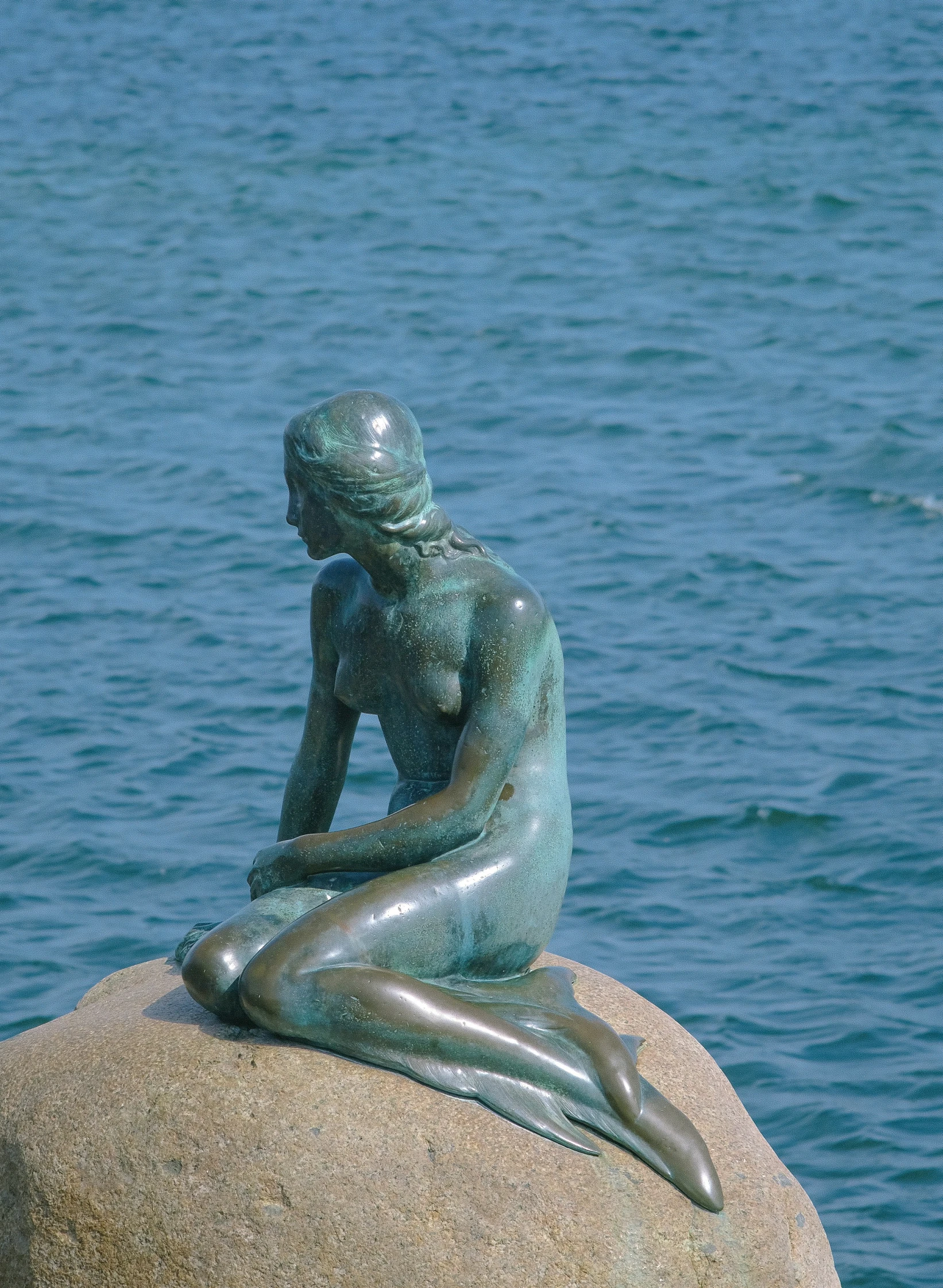 the little mermaid statue is sitting on the rock next to water