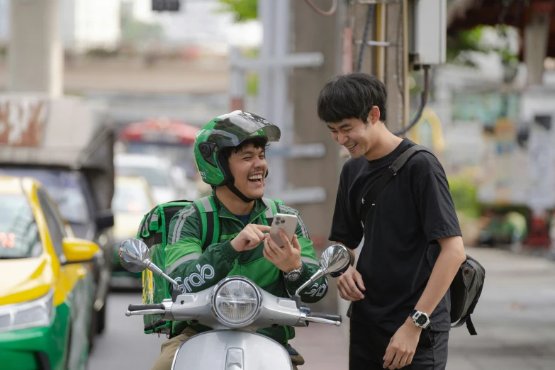 two men laughing while looking at a cell phone on a moped