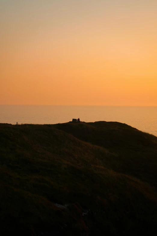 people sitting on the side of a hill with the sun setting over the water