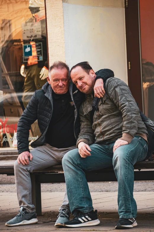 two men sitting on a bench together in front of a store