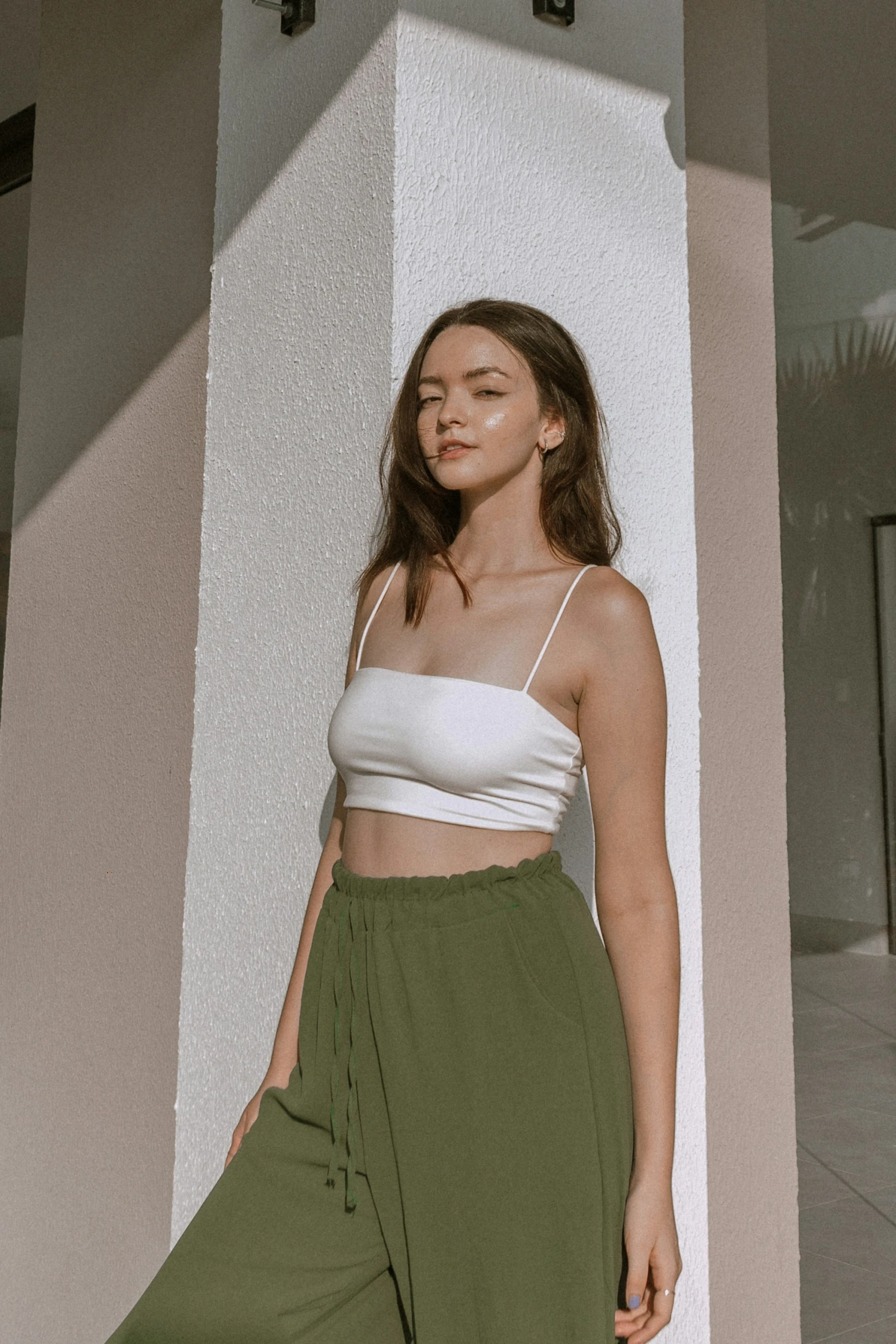 a woman stands against a wall wearing an open top and green pants