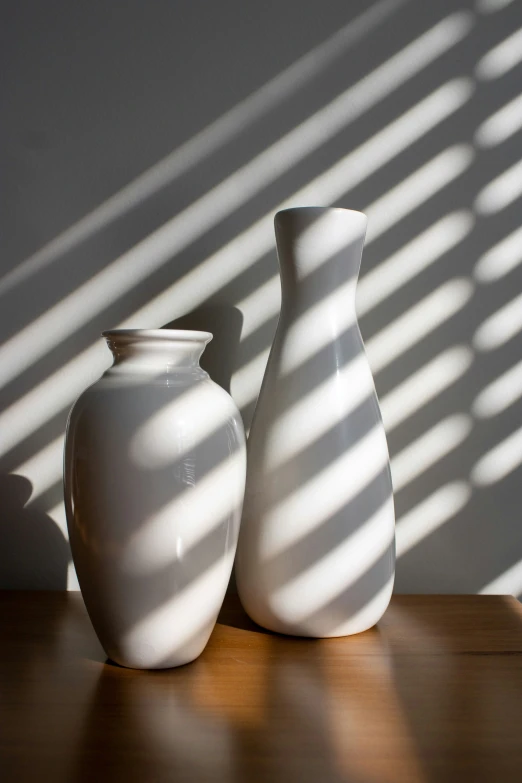 two white vases sitting on a table, one has a shadow cast on the wall behind it