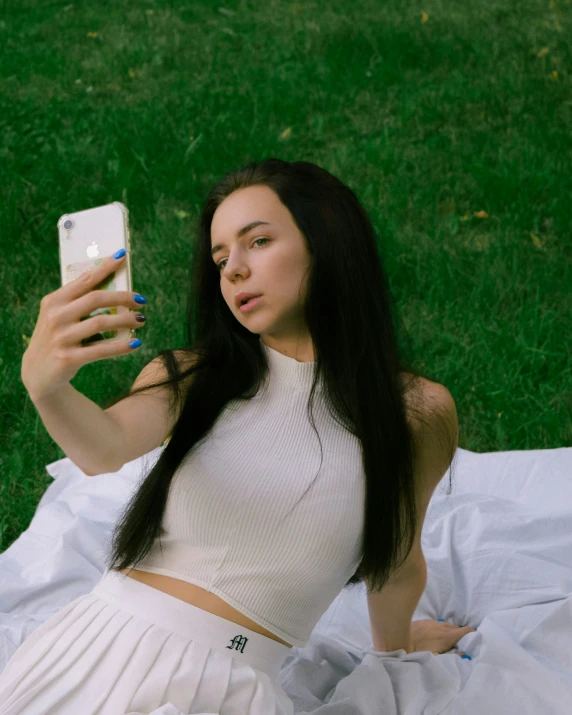 a girl in a white top is holding her cellphone while laying on the grass