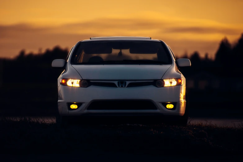 a white car with the lights on driving in front of a sunset