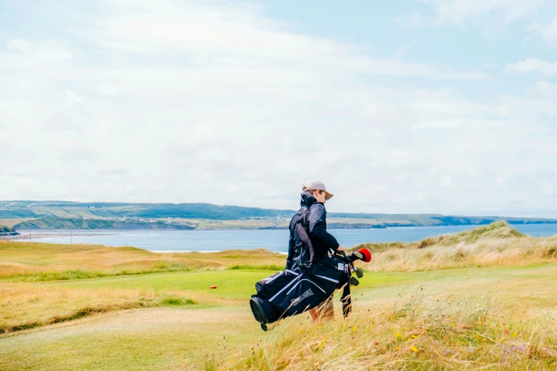 a man holding golf bag in grassy field with ocean and clouds