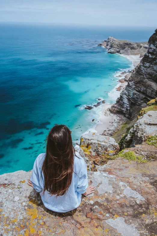 a woman is sitting on the edge of a cliff overlooking a clear ocean