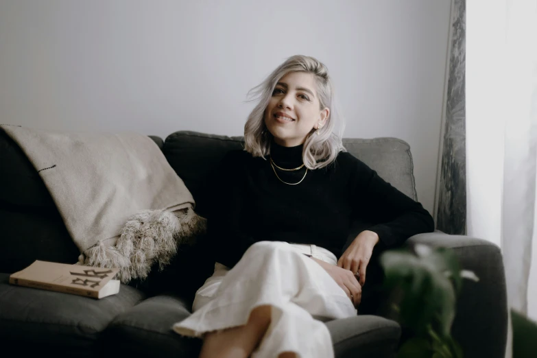 a woman with blond hair wearing a black sweater and white skirt sitting on a gray couch