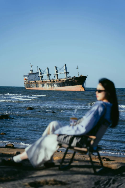 a person sitting on the edge of the beach watching a ship on a rocky shore