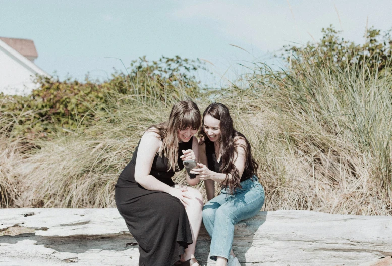 two woman sitting on a rock, taking pictures with a cellphone