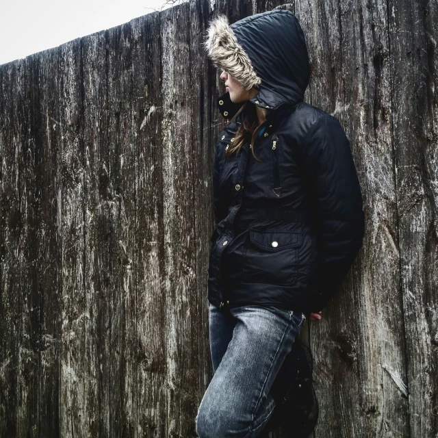 the person in hooded jacket leans against the wooden wall