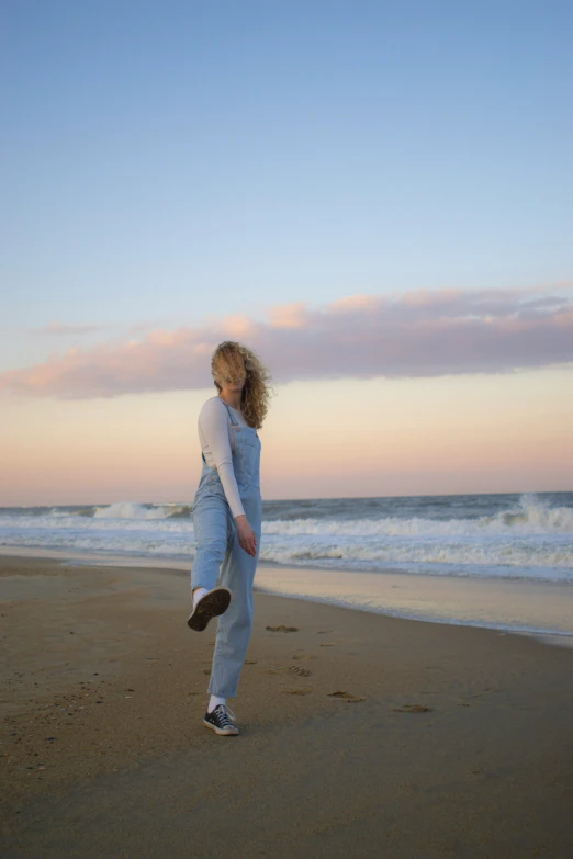 a girl stands on the beach in her overalls and she is facing away from the camera