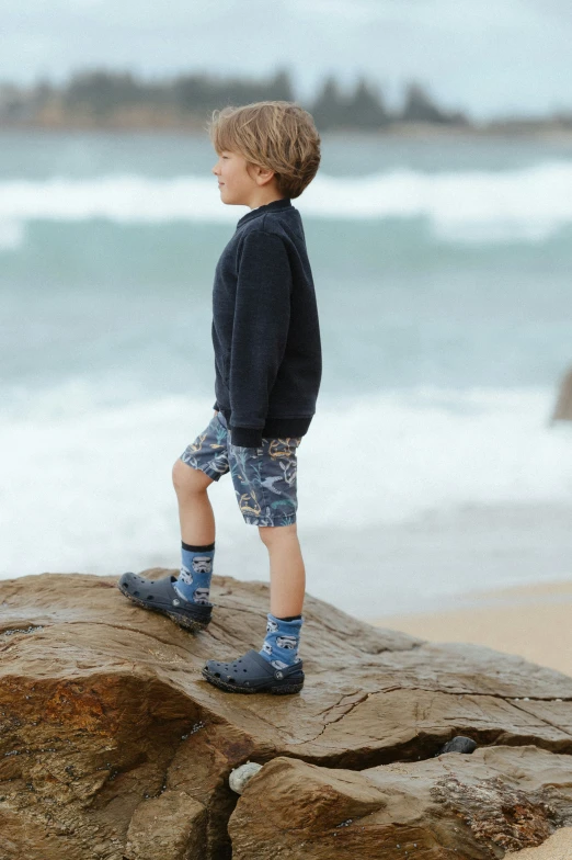 little boy in a pair of blue boots standing on a rock looking at the ocean