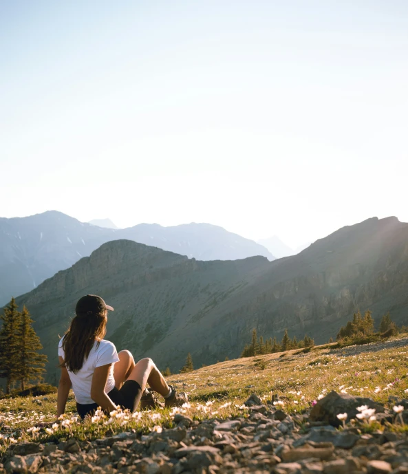 a woman sitting in the grass on a rocky hill with mountains behind her