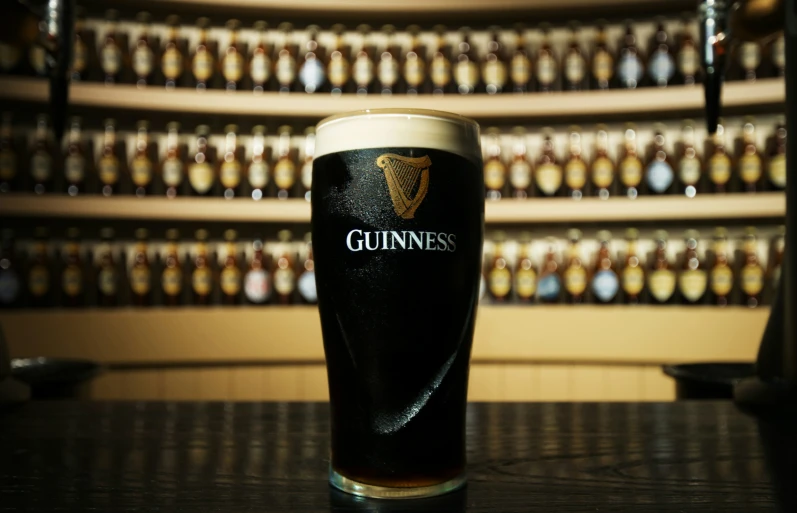 an irish pint sits on a counter in front of wall of guinness bottles