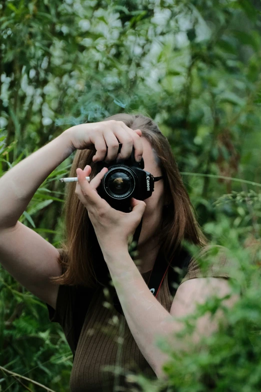 a woman holding a camera up to the side of her face in a forest