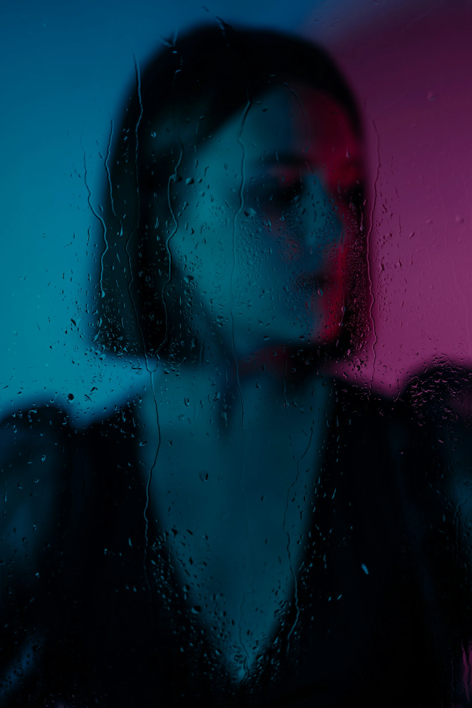 the woman has dark hair standing next to the window with water droplets on it