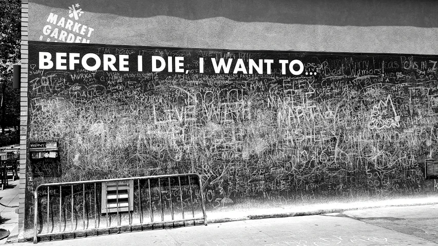 black and white pograph of people walking by wall with words that read before i die i want to