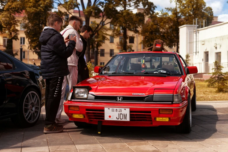 the people stand near a red car in front of several other cars